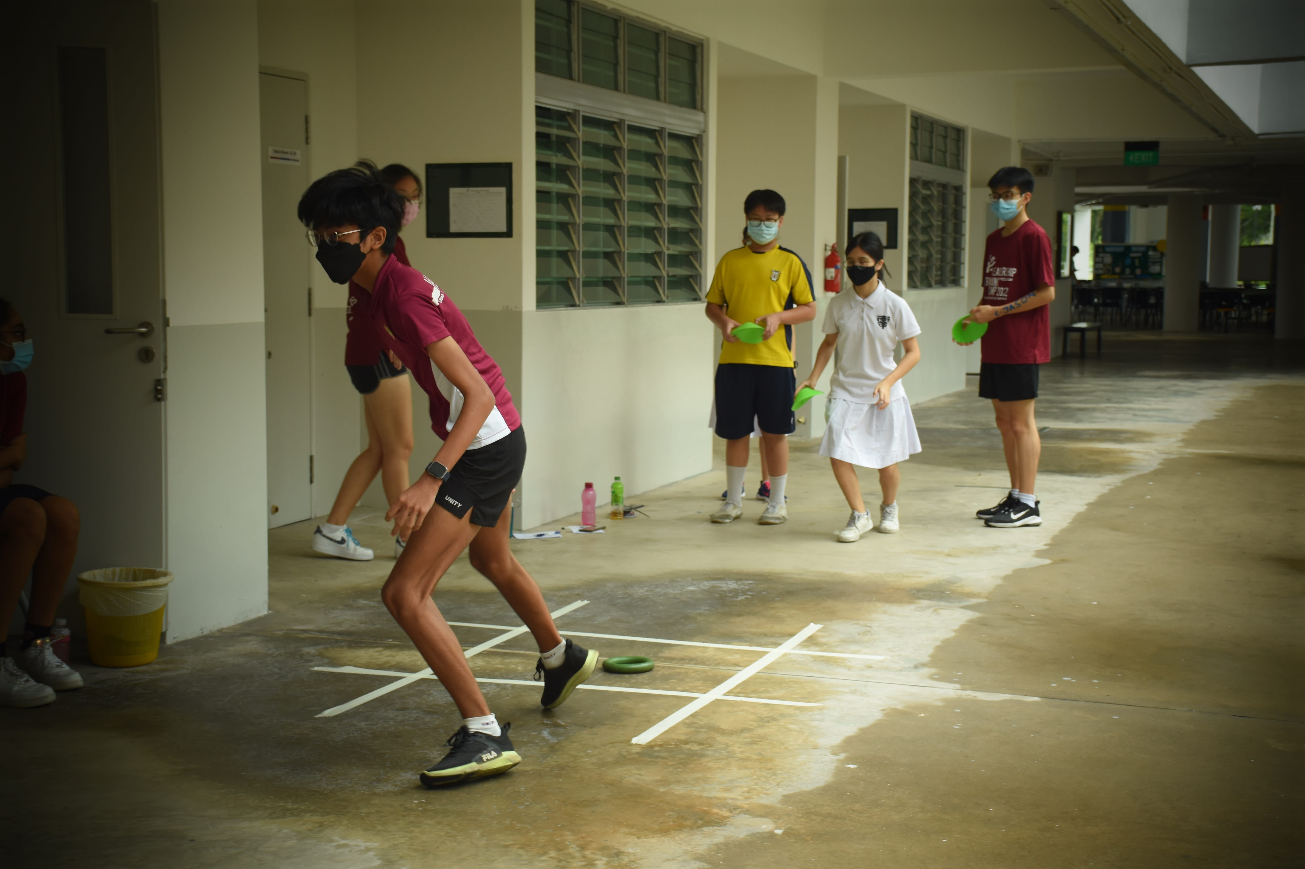 Students wearing face masks play a ring-toss game in a school corridor with taped targets on the floor.